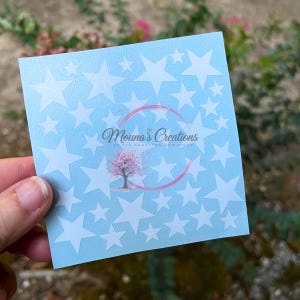 May include: A square light blue sheet featuring a repeating pattern of white stars in various sizes. Centered on the sheet is a circular logo containing the text Mouna's Creations with a small pink cherry blossom tree graphic below the text.