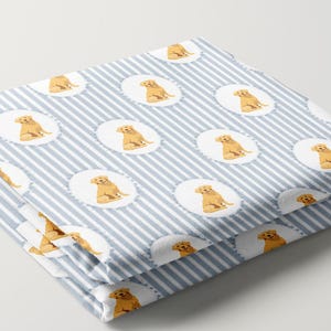 May include: Folded fabric with a light blue and white striped pattern. The fabric features repeating oval medallions with illustrations of golden retriever puppies. The text "Mockup view. Not a real view/size of fabric. See sizes on next photos." is visible.