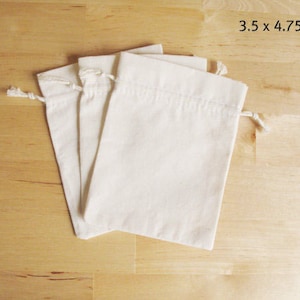 以下が含まれることがあります： オフホワイトの巾着袋が3つ、それぞれ約8.9 x 12.1 cmで、明るい木製の表面に配置されています。袋はプレーンな生地でできており、上部に紐で閉じるようになっています。ニュートラルな色とシンプルなデザインは、用途の広い製品を示唆しています。