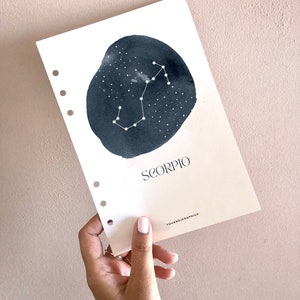 Puede incluir: Una tarjeta blanca con un círculo de acuarela negro que representa la constelación de Escorpio. El texto "SCORPIO" está impreso en negro debajo del círculo. El texto "YOUANDIGRAPHICS" está impreso en negro en la parte inferior de la tarjeta.