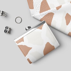 May include: A small, white and brown patterned coin purse with a silver clasp. The purse is lying on a white surface with silver rings nearby.