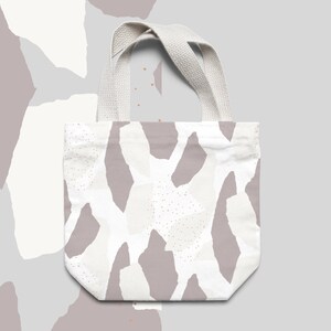 May include: A white tote bag with a pattern of abstract shapes in shades of gray and white. The bag has a white canvas strap.