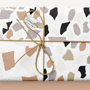 May include: A white gift box wrapped in paper with a terrazzo pattern in shades of brown, beige, gray, and black. The box is tied with a brown twine bow.