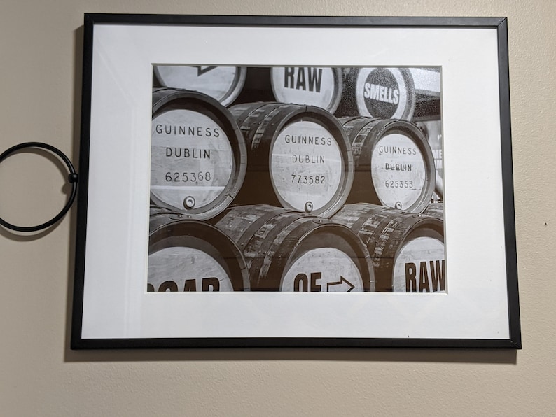 K&ouml;nnte beinhalten: Schwarz-Wei&szlig;-Druck in einem schwarzen Rahmen mit gestapelten Guinness-F&auml;ssern. Die F&auml;sser sind mit "Guinness Dublin" und Seriennummern beschriftet, sowie mit den W&ouml;rtern "RAW" und "SMELLS". Der Druck hat ein wei&szlig;es Passepartout.