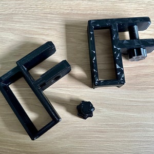 May include: Black 3D-printed C-clamps with a textured surface and a screw mechanism. One clamp is assembled, and the other is disassembled, with a separate knob. The clamps are designed to secure objects.
