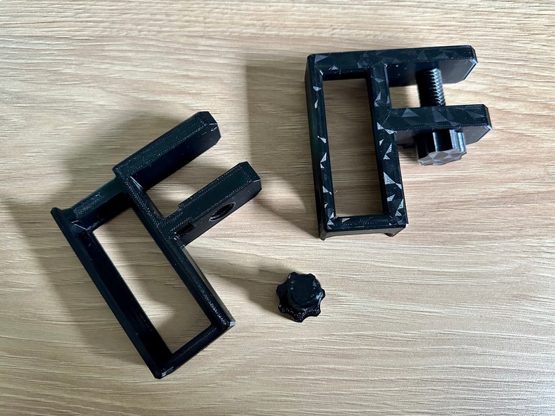 May include: Black 3D-printed C-clamps with a textured surface and a screw mechanism. One clamp is assembled, and the other is disassembled, with a separate knob. The clamps are designed to secure objects.
