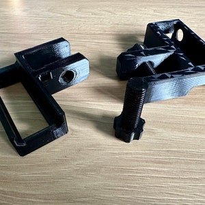 May include: Black 3D-printed mechanical components, including a bracket, a frame, and a bolt. The bracket has a rectangular opening and a threaded hole. The bolt has a hexagonal head. The components are designed for mechanical assembly.