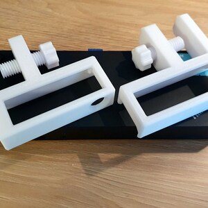 May include: Two white, C-shaped clamps with adjustable screws, resting on a black rectangular object. The clamps are made of a solid, opaque material and appear to be 3D-printed. The background is a light-colored wooden surface.