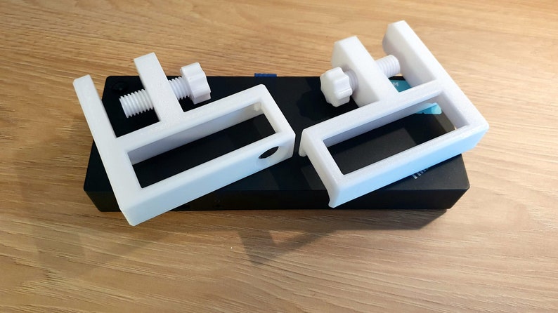 May include: Two white, C-shaped clamps with adjustable screws, resting on a black rectangular object. The clamps are made of a solid, opaque material and appear to be 3D-printed. The background is a light-colored wooden surface.