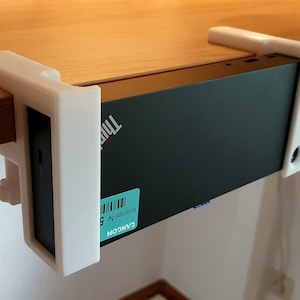May include: A black ThinkPad computer secured to a wooden surface with a white 3D-printed bracket. The bracket has a screw for tightening and a label with text and a barcode is visible on the computer.