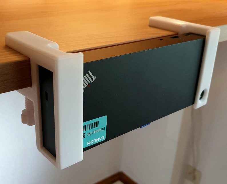 May include: A black ThinkPad computer secured to a wooden surface with a white 3D-printed bracket. The bracket has a screw for tightening and a label with text and a barcode is visible on the computer.