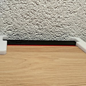 May include: A white 3D-printed phone stand with a black phone and red accent. The stand has a simple, modern design, and is placed on a wooden surface. The background is a textured white wall.