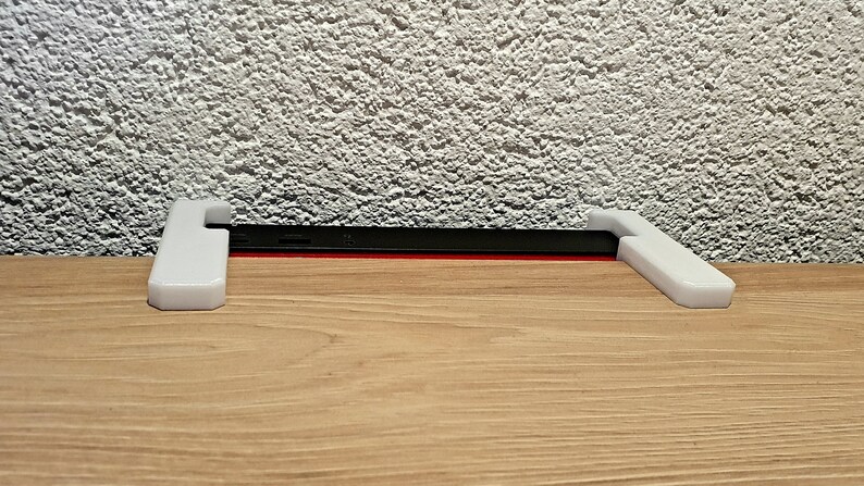 May include: A white 3D-printed phone stand with a black phone and red accent. The stand has a simple, modern design, and is placed on a wooden surface. The background is a textured white wall.