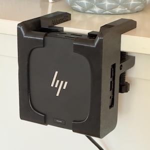 Op de afbeelding: Zwarte computerhouder met de tekst "Akkurat3D" en een HP-logo. De houder is met een klem aan een wit oppervlak bevestigd. De houder is ontworpen om een kleine computer vast te houden.