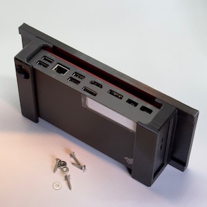 Op de afbeelding: Een zwart computer docking station met meerdere poorten, waaronder USB en HDMI, en een rode accentkleur. Het station is rechthoekig en heeft een strak ontwerp. Verschillende schroeven en ringen liggen in de buurt.