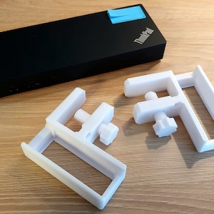 May include: Two white, 3D-printed C-clamps with adjustable screws and a black laptop with the ThinkPad logo and a blue sticky note. The clamps are designed to secure objects.