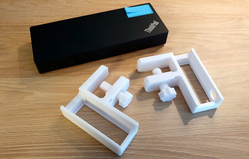 May include: Two white, 3D-printed C-clamps with adjustable screws and a black laptop with the ThinkPad logo and a blue sticky note. The clamps are designed to secure objects.