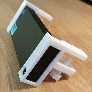 May include: A white 3D-printed clamp designed to hold a black rectangular object. The clamp has a screw mechanism for adjusting the grip and is resting on a wooden surface. The design includes a rectangular frame and supporting arms.