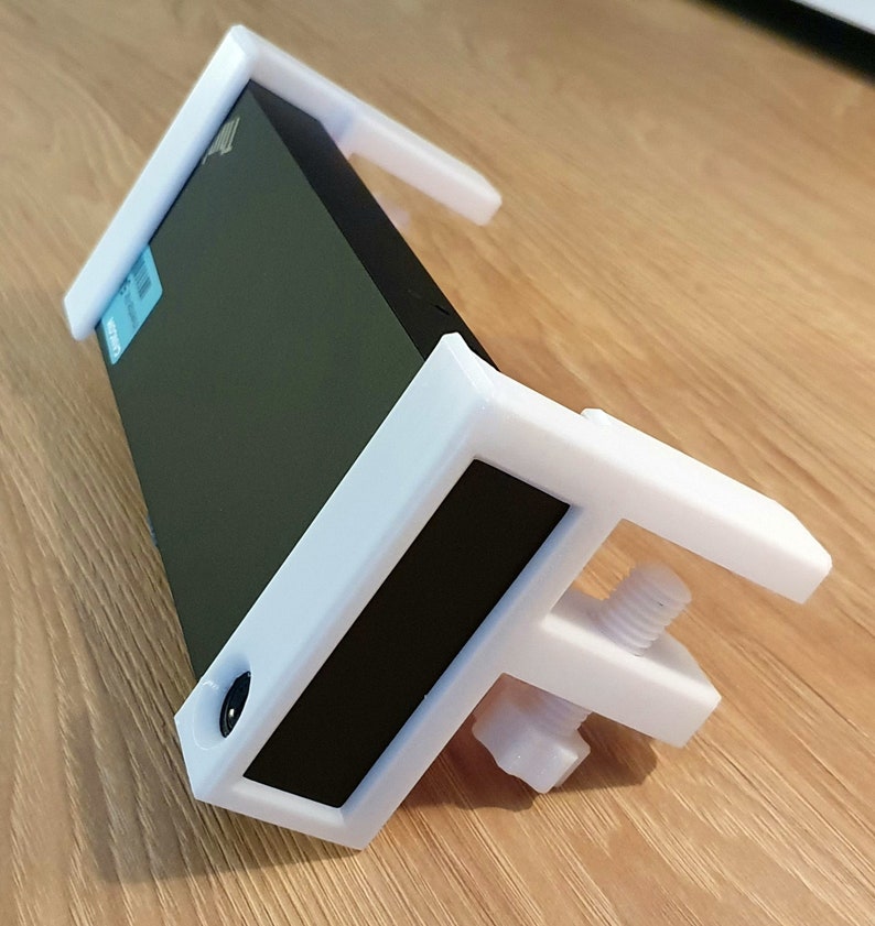 May include: A white 3D-printed clamp designed to hold a black rectangular object. The clamp has a screw mechanism for adjusting the grip and is resting on a wooden surface. The design includes a rectangular frame and supporting arms.