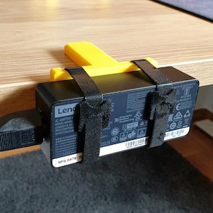 Op de afbeelding: Een zwarte Lenovo AC-adapter bevestigd onder een houten bureau met een gele plastic beugel en zwarte bandjes. De voedingskabel van de adapter is aangesloten en de fabricagedatum is zichtbaar.