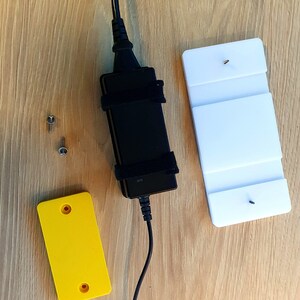 Könnte beinhalten: Ein schwarzes Netzteil mit schwarzem Kabel und Klettverschluss. Zwei Schrauben, ein gelbes Rechteck mit zwei Löchern und ein weißes Rechteck mit zwei Schrauben sind ebenfalls abgebildet. Alle Artikel liegen auf einer Holzoberfläche.