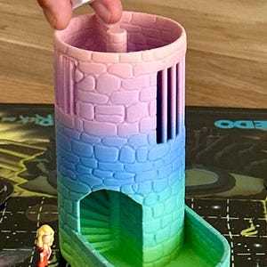 Könnte beinhalten: Ein mehrfarbiger Spielturm in Rosa, Blau und Grün mit einer Wendeltreppe und vertikalen Schlitzen. Zwei weiße Würfel mit schwarzen Punkten werden über dem Turm gehalten. Spielfiguren und ein Spielbrett sind sichtbar.