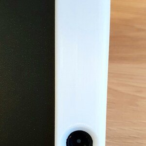 May include: A tall, white rectangular object with a black circular component at the bottom. The object is positioned next to a black, textured surface. The background is a light brown wooden surface.