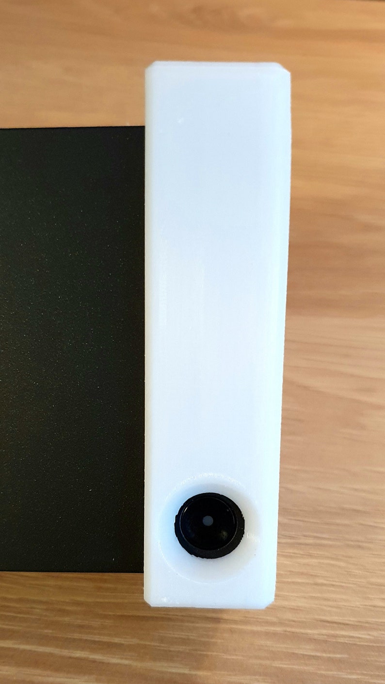 May include: A tall, white rectangular object with a black circular component at the bottom. The object is positioned next to a black, textured surface. The background is a light brown wooden surface.
