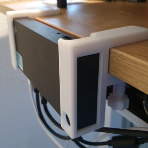 May include: A white, 3D-printed desk mount securely holds a black electronic device. The mount clamps onto a wooden desk, with a white screw for adjustment. Cables are visible below the device.