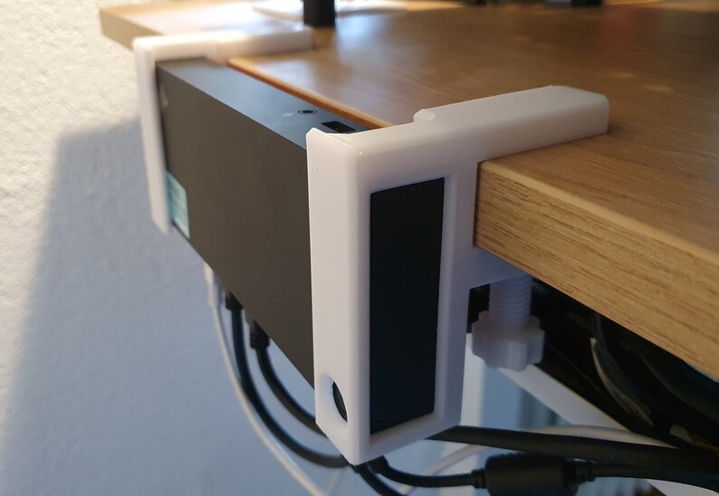 May include: A white, 3D-printed desk mount securely holds a black electronic device. The mount clamps onto a wooden desk, with a white screw for adjustment. Cables are visible below the device.