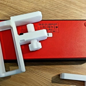 May include: White plastic mounting bracket with a threaded adjustment screw, positioned over a red rectangular device with black trim and red feet. The bracket is designed to hold or secure an item. The device has a label with text and symbols.