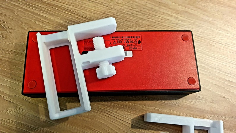 May include: White plastic mounting bracket with a threaded adjustment screw, positioned over a red rectangular device with black trim and red feet. The bracket is designed to hold or secure an item. The device has a label with text and symbols.