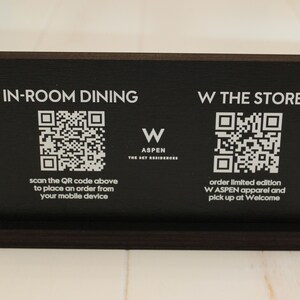 K&ouml;nnte beinhalten: Schwarzes Schild mit wei&szlig;em Text und QR-Codes. Das Schild lautet "IN-ROOM DINING" mit einem QR-Code und dem Text "Scannen Sie den QR-Code oben, um eine Bestellung von Ihrem Mobilger&auml;t aus aufzugeben". Das Schild lautet auch "W THE STORE" mit einem QR-Code und dem Text "Bestellen Sie limitierte W ASPEN-Kleidung und holen Sie sie bei Welcome ab".