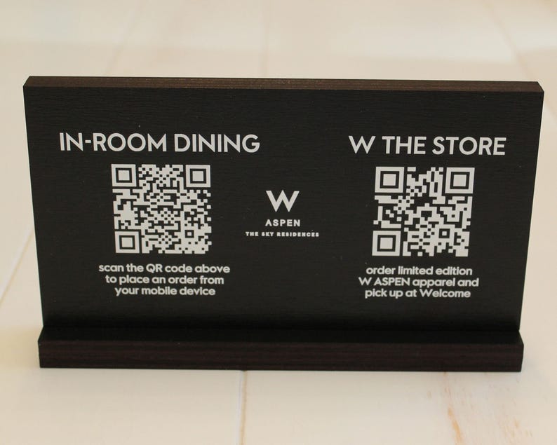 K&ouml;nnte beinhalten: Schwarzes Schild mit wei&szlig;em Text und QR-Codes. Das Schild lautet "IN-ROOM DINING" mit einem QR-Code und dem Text "Scannen Sie den QR-Code oben, um eine Bestellung von Ihrem Mobilger&auml;t aus aufzugeben". Das Schild lautet auch "W THE STORE" mit einem QR-Code und dem Text "Bestellen Sie limitierte W ASPEN-Kleidung und holen Sie sie bei Welcome ab".