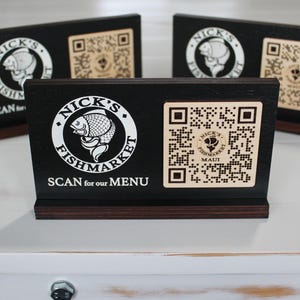 K&ouml;nnte beinhalten: Schwarzes Holzschild mit einem wei&szlig;en Logo, auf dem "Nick's Fishmarket Maui" steht, und einem QR-Code. Das Schild sagt "Scan for our menu".