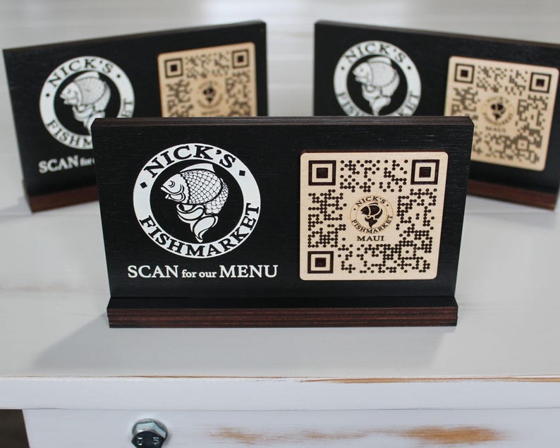 K&ouml;nnte beinhalten: Schwarzes Holzschild mit einem wei&szlig;en Logo, auf dem "Nick's Fishmarket Maui" steht, und einem QR-Code. Das Schild sagt "Scan for our menu".