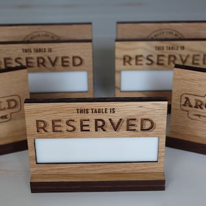 Può includere: Un set di segnatavolo rettangolari in legno con il testo inciso THIS TABLE IS RESERVED. Ogni segnatavolo presenta una finitura in legno chiaro, una base marrone scuro e un inserto rettangolare bianco per aggiungere testo personalizzato.