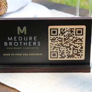 K&ouml;nnte beinhalten: Ein schwarzes Holzschild mit einem goldenen M, dem Text "Medure Brothers Culinary Concepts" und dem Text "Scan to view our concepts". Rechts auf dem Schild befindet sich ein goldener QR-Code.