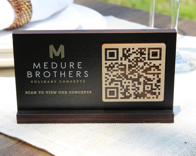 K&ouml;nnte beinhalten: Ein schwarzes Holzschild mit einem goldenen M, dem Text "Medure Brothers Culinary Concepts" und dem Text "Scan to view our concepts". Rechts auf dem Schild befindet sich ein goldener QR-Code.