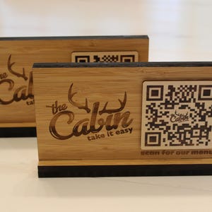 K&ouml;nnte beinhalten: Zwei Holzschilder mit schwarzem Rand und einem QR-Code. Die Schilder sind mit dem Text "The Cabin take it easy" und einem stilisierten Bild von Geweih verziert. Der Text "scan for our menu" ist ebenfalls auf dem Schild graviert.