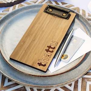 May include: A rectangular bamboo clipboard with a dark metal clip at the top and the word "thattu" laser-engraved on the front. A silver ballpoint pen and a gray credit card are placed on top of a white paper sheet resting on the clipboard.
