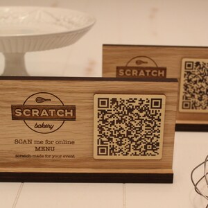 K&ouml;nnte beinhalten: Ein Schild aus Holz mit einer braunen Basis und einer hellbraunen Oberseite. Das Schild tr&auml;gt ein Logo f&uuml;r "Scratch Bakery" mit einem Schneebesen und dem Text "SCAN me for online MENU scratch-made for your event". Ein QR-Code befindet sich auf dem Schild.