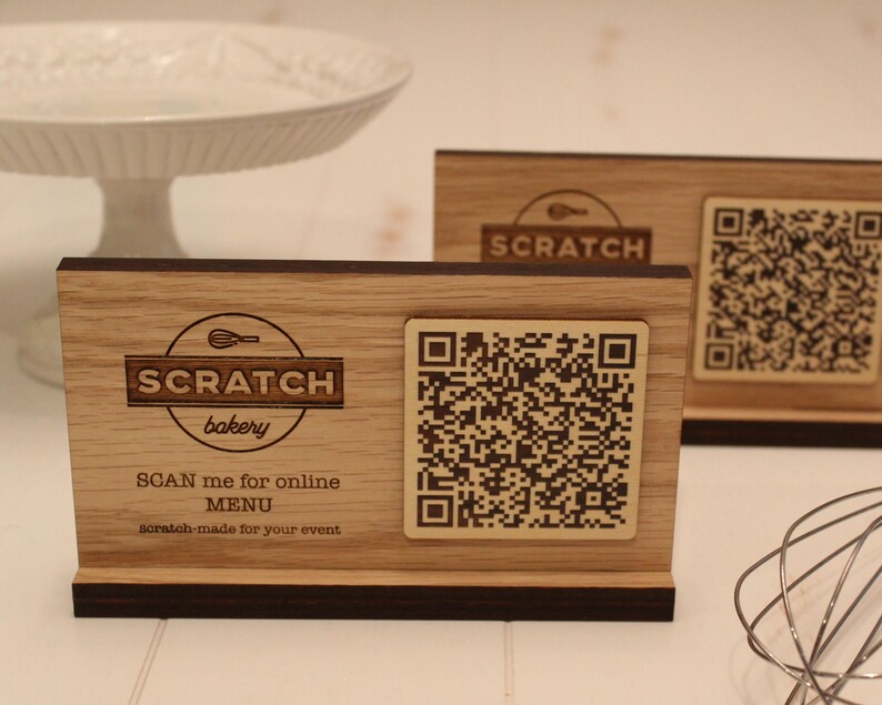 K&ouml;nnte beinhalten: Ein Schild aus Holz mit einer braunen Basis und einer hellbraunen Oberseite. Das Schild tr&auml;gt ein Logo f&uuml;r "Scratch Bakery" mit einem Schneebesen und dem Text "SCAN me for online MENU scratch-made for your event". Ein QR-Code befindet sich auf dem Schild.
