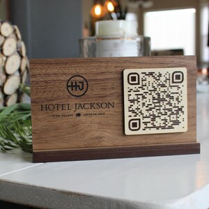 K&ouml;nnte beinhalten: Ein Schild aus Holz mit einer dunkelbraunen Oberfl&auml;che und einem schwarzen, gravierten Logo mit der Aufschrift "Hotel Jackson" sowie dem Text "Town Square" und "Jackson Hole" darunter. Das Schild hat au&szlig;erdem einen schwarz-wei&szlig;en QR-Code.