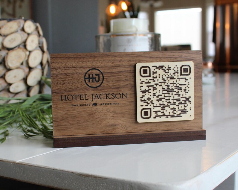 K&ouml;nnte beinhalten: Ein Schild aus Holz mit einer dunkelbraunen Oberfl&auml;che und einem schwarzen, gravierten Logo mit der Aufschrift "Hotel Jackson" sowie dem Text "Town Square" und "Jackson Hole" darunter. Das Schild hat au&szlig;erdem einen schwarz-wei&szlig;en QR-Code.
