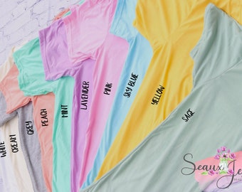 Pode incluir: Uma coleção de camisetas de manga curta em várias cores, incluindo branco, creme, cinza, pêssego, menta, lavanda, rosa, azul celeste, amarelo e sálvia. Cada camiseta é rotulada com o nome da cor correspondente em texto preto no tecido.