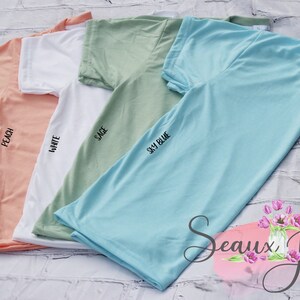 May include: Four short-sleeved t-shirts in peach, white, sage green, and sky blue. Each shirt is labeled with its respective color name. A logo for Seaux Jessi's Beautiful Blanks featuring a vase of pink tulips is in the bottom right corner.