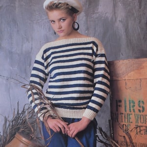 Può includere: Una donna indossa un maglione a righe bianche e blu con un berretto bianco e grandi orecchini a cerchio. È in piedi davanti a una cassa di legno con la scritta "FIRST IN THE WORLD OF TEA" stampata su di essa.