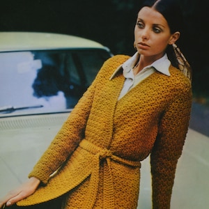 May include: A woman wearing a mustard yellow crocheted cardigan with a self-tie belt. The cardigan has a V-neck and long sleeves.