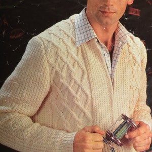 May include: A cream-colored knitted cardigan with a cable knit pattern and a v-neck. The cardigan is buttoned up and the person is holding a fishing reel.